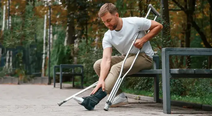 Hombre joven con pierna lesionada usa muletas, sentado en un banco.
